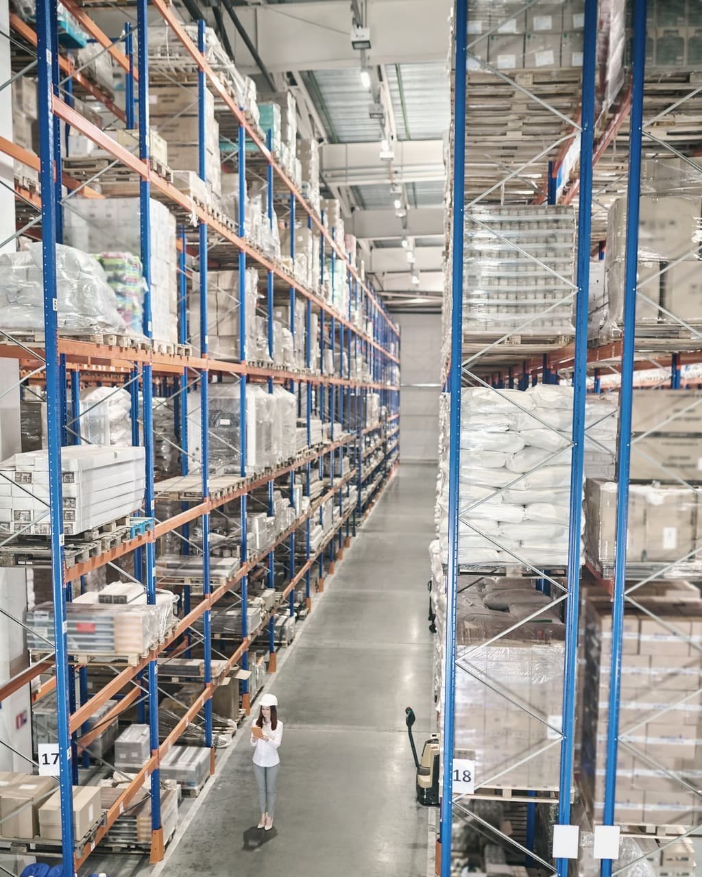 A distribution warehouse aisle lined with high racks of stock, with a worker checking inventory on a tablet