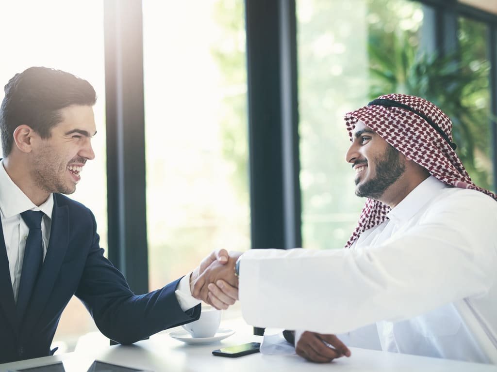 Two business partners shaking hands across a desk in a bright office