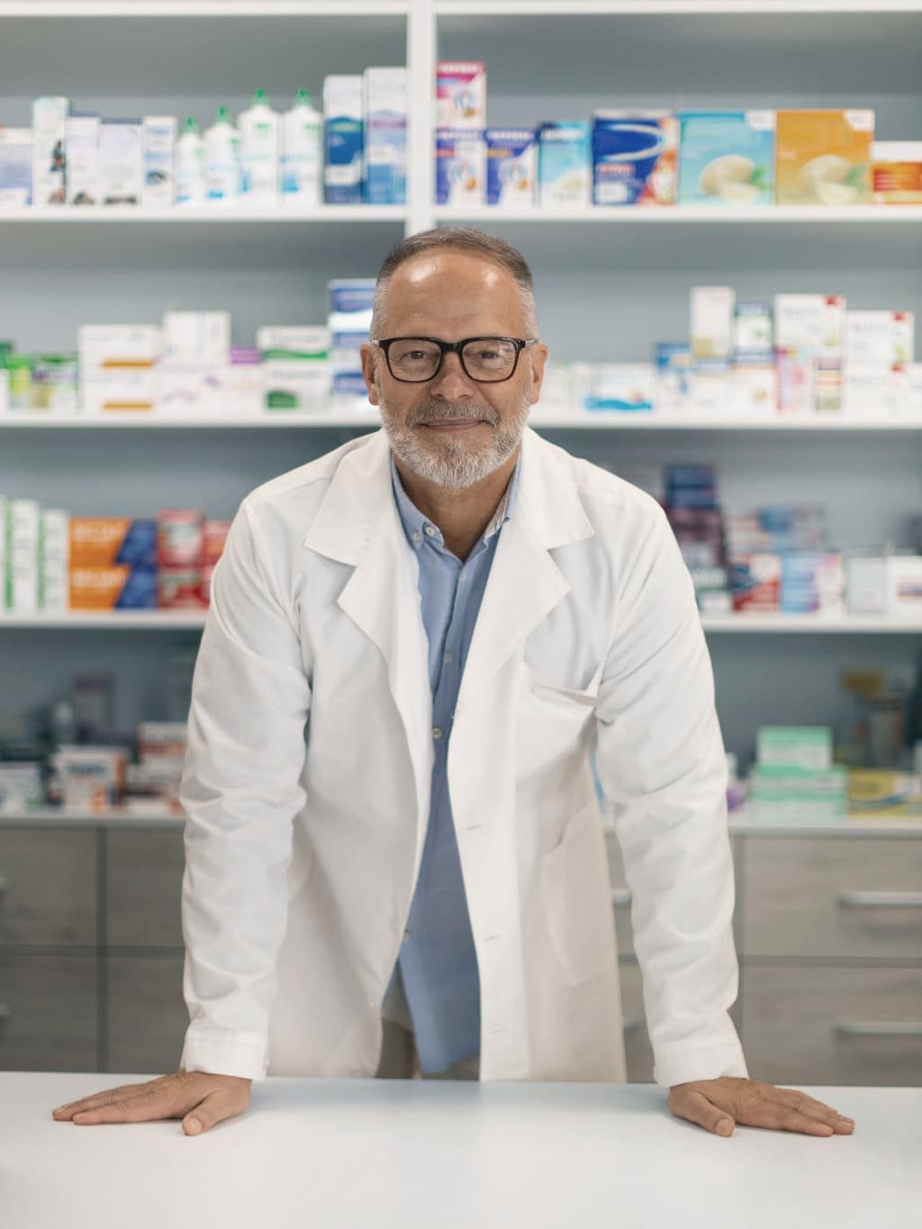 A smiling pharmacist leaning on the counter of a well-stocked pharmacy